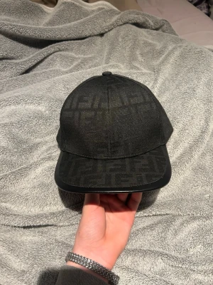 Svart keps från Fendi med logomönster - Snygg svart keps från Fendi med det klassiska FF-logomönstret över hela kepsen. Justerbar rem baktill och detaljer i svart tyg. Perfekt accessoar för dig som gillar exklusiv streetwear och vill sticka ut med en lyxig touch.