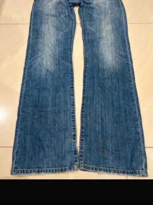Blå bootcut jeans med slitningar - Säljer ett par blå bootcut jeans med slitningar och ljusa tvättade partier på låren. Jeansen har klassisk femficksdesign och är tillverkade i denim. Perfekta för dig som gillar en avslappnad och retro look.