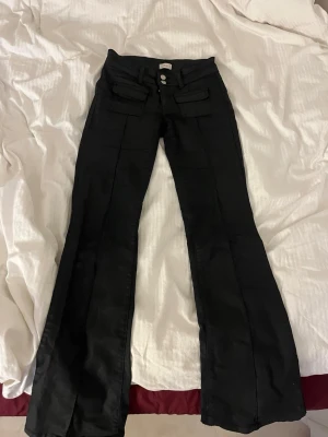 Svarta bootcut byxor med fickor - Snygga svarta byxor med bootcut passform och dubbla knappar i midjan. Byxorna har framfickor med lock och markerade sömmar längs benen. Perfekta för dig som gillar en trendig och lite retro look.