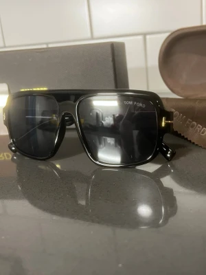 Solglasögon  - Stilrena svarta solglasögon från Tom Ford med rektangulära glas och bred båge. Snygga gulddetaljer vid gångjärnen och mörka linser. Kommer med originalfodral och putsduk. Perfekt accessoar för en trendig look.