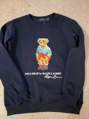  Polo Bear hoodie Ralph Lauren - Svart hoodie från Ralph Lauren med ikoniska Polo Bear-trycket framtill. Klassisk passform med ribbade muddar och rund hals. Perfekt för dig som gillar streetstyle och vill sticka ut med en cool björn på bröstet.