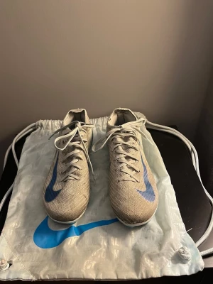 Nike Mercurial Zoom fotbollsskor grå/blå - Nike Mercurial Zoom fotbollsskor i grått med blå detaljer och snörning. Skorna har en smal passform och är tillverkade i syntetmaterial med mönstrad ovandel för bättre bollkontroll. Perfekta för dig som vill ha lättviktsskor med bra grepp på planen.