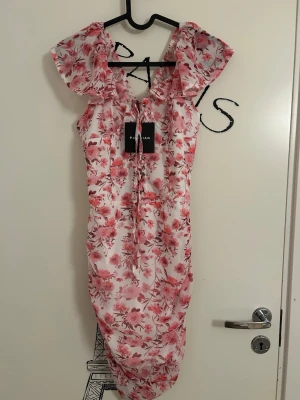 Parisian Floral Bardot Mini Dress - Söt blommig klänning i vitt och rosa med snörning framtill och dragkedja därbak. Från märket Parisian i storlek XS och är köpt på Nelly. Klänningen  har drapering och volangärm. Aldrig använd och har lappen kvar. Helt slutsåld klänning på Nellys hemsida. 