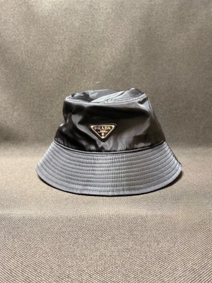 Svart bucket hat från Prada - Stilren svart bucket hat från Prada med klassisk triangellogga framtill. Hatten är tillverkad i glansig syntet och har en mjuk, nedvikt brätte. Perfekt accessoar för en trendig look.