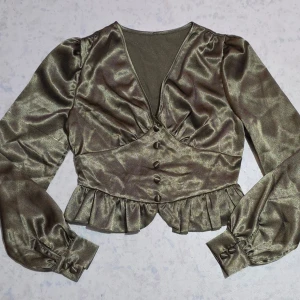  glansig blus med volang - Stilren blus i glansigt material med v-ringning, långa puffärmar och knappar framtill. Bl soppen har volang nedtill och markerad midja för en snygg siluett. Perfekt för dig som gillar trendiga detaljer och en feminin look.