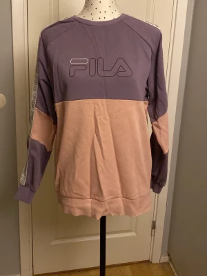 Lila och rosa sweatshirt från FILA - Snygg sweatshirt från FILA i lila och ljusrosa färgblock. Tröjan har stora FILA-loggor på bröstet och längs ärmarna, samt ribbade muddar vid ärmslut och nederkant. Oversized passform och mjukt material som känns skönt mot huden.