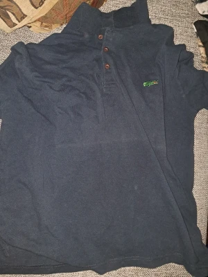 Mörkblå piké från Lacoste - Klassisk mörkblå pikétröja från Lacoste med den ikoniska gröna krokodilen broderad på bröstet. Tröjan har krage, knappar framtill och är tillverkad i bomull för en skön känsla. Perfekt för en avslappnad men ändå stilren look.