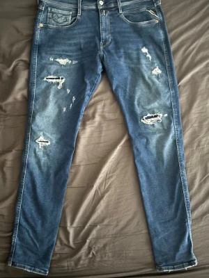 Blå Replay Anbass jeans med slitningar - Säljer ett par blå Replay Anbass jeans med snygga slitningar och distressed detaljer på benen. Midja 34 och längd 32 