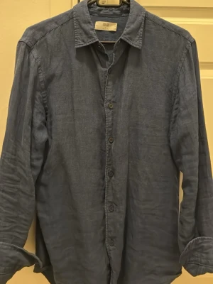 Blå linneskjorta från Uniqlo - Säljer en blå skjorta från Uniqlo i mjukt linne. Skjortan har klassisk krage, knappar hela vägen och långa ärmar. Perfekt för dig som gillar en avslappnad men ändå snygg look. Fräsch till sommaren!