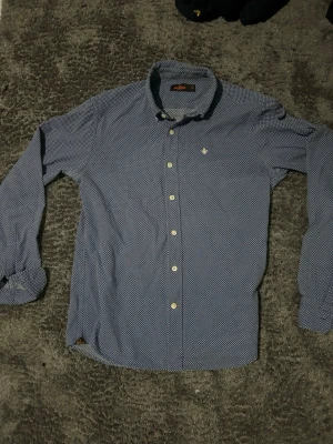 Blå mönstrad skjorta från Morris - Snygg blå skjorta från Morris med diskret prickigt mönster och klassisk krage. Skjortan har vita knappar framtill och en liten broderad logga på bröstet. Tillverkad i bomull och har lång ärm för en stilren look.