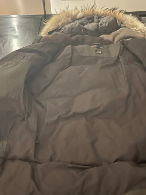 Mörkgrön dunjacka med fuskpäls från candiens  - Snygg mörkgrön dunjacka med huva och fuskpäls runt kanten. Jackan har en quiltad design och dragsko i midjan för bättre passform. Perfekt för kalla dagar och har en klassisk look som funkar till det mesta.