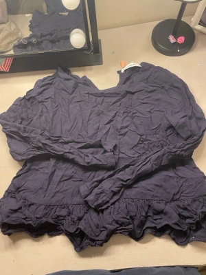 Mörkblå volangblus med lång ärm - Mörkblå blus med volangkant nedtill och långa ärmar. Blusen har en avslappnad passform och rund halsringning. Materialet ser ut att vara mjukt och lätt, perfekt för en trendig och bekväm look.