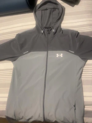 Grå vindjacka från Under Armour - Snygg tvåtonad grå vindjacka från Under Armour med huva och hel dragkedja framtill. Jackan har lång ärm, justerbar huva med snörning och logga på bröstet. Perfekt för blåsiga dagar och sportiga outfits.