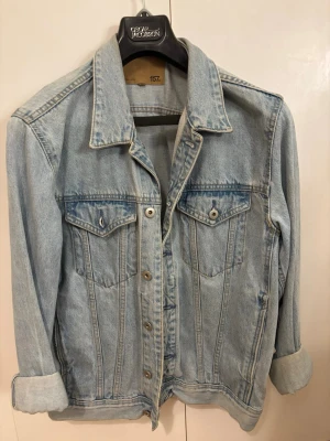 Ljusblå jeansjacka från 157 - Snygg ljusblå jeansjacka från 157 med klassisk krage, knäppning framtill och två bröstfickor med lock. Jackan har långa ärmar och en avslappnad passform, perfekt att slänga över en hoodie eller t-shirt.