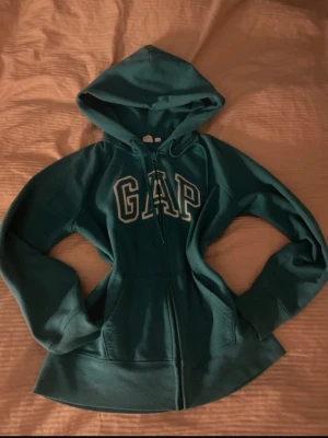 Turkos hoodie med dragkedja från GAP - Snygg turkos hoodie från GAP med dragkedja framtill och stor huva. Klassisk logga i vitt på bröstet och två fickor fram. Perfekt för en avslappnad och cool stil. Tillverkad i mjukt bomullsmaterial som känns skönt mot huden.