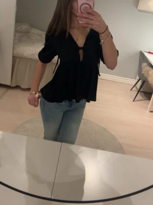 Svart v-ringad topp med kort ärm - Säljer en svart topp med v-ringning och korta puffärmar. Toppen har en liten knytning framtill och ett löst, flowy fall. Perfekt till jeans eller kjol för en trendig look. Materialet ser ut att vara mjukt och bekvämt, troligen bomull eller viskos.