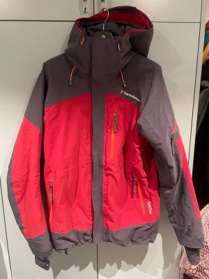 Röd och grå vindjacka Peak Performance - Snygg röd och mörkgrå vindjacka från Peak Performance med huva och flera dragkedjeförsedda fickor. Jackan har justerbara snören vid huvan och detaljer i orange. Perfekt för blåsiga dagar och outdoor-äventyr. Har en lagning på luvan som inte påverkar mer än de kosmetiska 