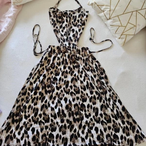 Leopardmönstrad klänning med knytband - En leopardmönstrad klänning med halterneck och öppen rygg. Klänningen har en tight passform( bredd måtten 35-39 cm , stretchig) upptill och en vid kjol. Kjolen är 65cm lång. Klänningen är ärmlös och har knytband som kan knytas på olika sätt. Perfekt för sommaren  Passar till stl S, även M, är lite stretchig.