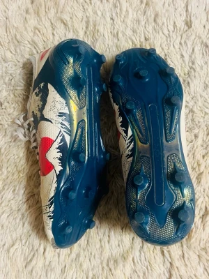 Vita och blå fotbollsskor med vågmönster - Unika fotbollsskor med vit ovandel, blå sula och coolt vågmönster i blått och rött. Skorna har snörning och är tillverkade i syntetmaterial. Perfekta för dig som vill sticka ut på planen med stilrena detaljer och färgstark design.