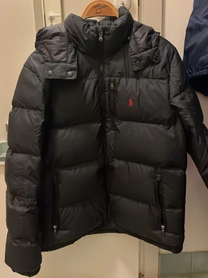 Svart dunjacka från Ralph Lauren - Svart puffer dunjacka från Ralph Lauren med avtagbar huva och dragkedja framtill. Jackan har två fickor med dragkedja och den klassiska röda Polo-loggan broderad på bröstet. Perfekt för kalla dagar.