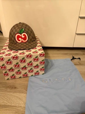 Gucci GG Supreme keps med äpple - Säljer en exklusiv Gucci keps i GG Supreme canvas med ikoniskt körsbärsmotiv framtill. Kepsen är brun med Guccis klassiska monogrammönster och har justerbart rött spänne bak. Kommer med originalbox och dustbag. Perfekt för dig som vill sticka ut med lyxig streetstyle. Som ny!!! Den passar även M!!
