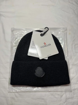 Moncler black label beanie  - Snygg svart ribbstickad mössa från Moncler med uppvikt kant och Moncler-logga framtill. Klassisk och stilren design som passar perfekt till höst och vinter. Tillverkad i mjukt material för extra komfort.