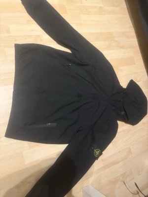 Svart vindjacka från Stone Island - Svart vindjacka från Stone Island med huva och dragkedja framtill. Jackan har två fickor med dragkedja och den klassiska Stone Island-loggan på vänster ärm. Perfekt för blåsiga dagar och stilren streetwear-look.