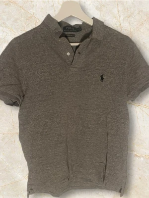 Grå pikétröja från Polo Ralph Lauren - Klassisk grå pikétröja från Polo Ralph Lauren med broderad logga på bröstet. Tröjan har korta ärmar, krage och knappar framtill. Modellen är custom slim fit och passar perfekt till en avslappnad men stilren look.