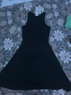 Svart ärmlös klänning från H&M - Stilren svart klänning från H&M i storlek XS. Klänningen är ärmlös med rund halsringning och har en utsvängd kjol som ger en klassisk siluett. Perfekt för dig som gillar enkel och tidlös design.