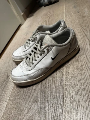 Vita Nike sneakers  storlek 42 -  vita sneakers från Nike med klassisk svart swoosh på sidan och broderad Nike-logga på hälen. Skorna har perforerad tå, grå snörning och en låg profil. Ovandelen är i läder och insidan är vadderad för extra komfort.