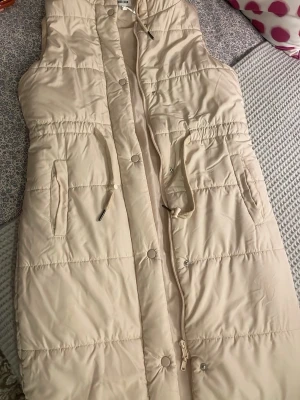 Lång beige vadderad väst med huva - Snygg lång beige vadderad väst med huva och dragsko i midjan. Västen har två fickor med dragkedja och stängs med både dragkedja och tryckknappar. Perfekt för lager på lager och en chill look.