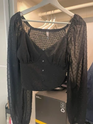 Svart transparent topp med knappar - Trendig svart topp med transparenta, prickiga ärmar och v-ringad hals. Croppad modell med knappar framtill och smockad rygg. Lätt och luftig, perfekt att styla med höga jeans eller kjol.