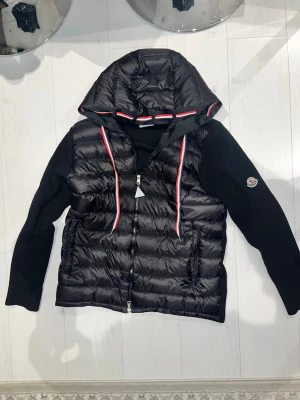 Svart Moncler cardigan  - Snygg svart dunjacka från Moncler med huva och dragkedja framtill. Jackan har stickade ärmar och Moncler-logga på ena ärmen. Fodret är quiltat och jackan har detaljer i rött, vitt och blått längs huvan och dragkedjan. Perfekt för kyliga dagar.