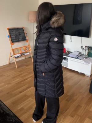 Svart lång dunjacka från Moncler - Snygg svart lång dunjacka från Moncler med huva och fuskpäls. Jackan har quiltad design, dragkedja framtill och Moncler-logga på ärmen. Perfekt för kalla dagar och ger en stilren look.