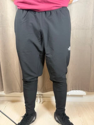 Svarta mjukisbyxor från The North Face - Svarta mjukisbyxor från The North Face med smal passform och elastisk midja. Byxorna har en stilren look med logga på låret och är gjorda i ett mjukt syntetmaterial som känns skönt mot huden. Perfekta för chill eller träning.