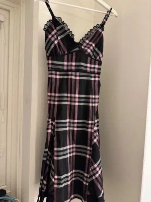 Rutig svart och rosa klänning - Snygg kort klänning med svart, rosa och grått rutmönster. Klänningen har smala axelband, spetsdetaljer vid urringningen och omlott upptill. Perfekt för dig som gillar en edgy och söt stil.