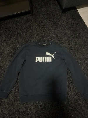 Svart stickad tröja från Puma - Snygg svart stickad tröja från Puma med stort vitt logotryck på bröstet. Tröjan har rund halsringning, långa ärmar och ribbade muddar vid ärmslut och nederkant. Perfekt för en avslappnad och sportig stil.
