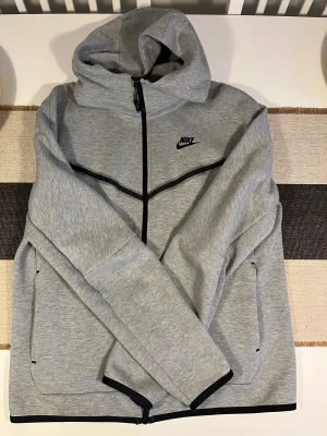 Nike Tech Grå hoodie  med dragkedja - Snygg grå hoodie från Nike med svart logga på bröstet och svarta detaljer längs dragkedjan och fickorna. Tröjan har huva, långa ärmar och praktisk hel dragkedja framtill. Perfekt för chill eller träning, med mjukt material som känns skönt mot huden. Unisex. Använd bara 2-3 gånger.