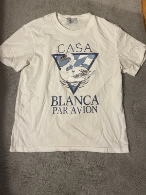 Casablanca Par Avion T-shiet - Säljer en vit t-shirt från Casablanca med tryck av en svan och texten 'Casa Blanca Par Avion' i blått framtill. Klassisk passform och rund hals. Perfekt för dig som gillar stilrena plagg med cool grafik. Denna tröja är rätt så sällsynt då den ej förekommer ofta, jag väljer dock att sälja den då den ej kommer till användning lika ofta längre, som man ser på skrynklet😅. Priset är ej hugget i sten och vid frågor så är det bara att ställa!