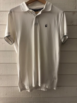 Vit pikétröja från Sail Racing - Klassisk vit pikétröja från Sail Racing med korta ärmar och liten svart logga på bröstet. Tröjan har krage och knappar vid halsen samt diskreta svarta detaljer vid ärmsluten. Perfekt till en clean och stilren look.