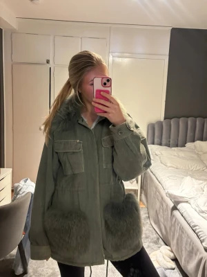 Grön parkajacka med fuskpälsdetaljer - Storlek S/M Custommade jacka!!!Snygg grön parkajacka med stora fickor och mjuka fuskpälsdetaljer vid krage och fickor. Jackan har en loose passform, dragkedja framtill och praktiska bröstfickor med lock. Perfekt för kyligare dagar.
