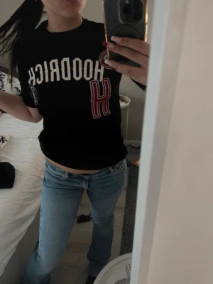 Svart t-shirt från Hoodrich - Svart t-shirt från Hoodrich med vita och röda tryck och patchar framtill. Stor vit text med märkesnamnet och flera detaljerade emblem. Klassisk passform och rund halsringning. Perfekt för en streetwear-look.