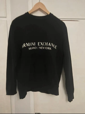 Svart sweatshirt Armani Exchange - Svart sweatshirt från Armani Exchange med vit logga och texten 'Milano / New York' på bröstet. Klassisk rund halsringning och långa ärmar. Perfekt för en stilren och trendig look.