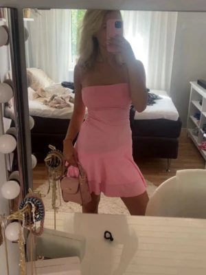 Axelbandslös rosa kortklänning - Söt axelbandslös klänning i ljusrosa med volangdetalj nertill. Klänningen har en figurnära passform och är sydd i ett mjukt material som ger en bekväm känsla. Perfekt för sommarens varma dagar och tillfällen när du vill sticka ut lite extra.