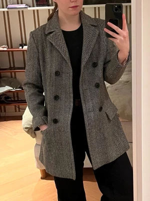 Grå chic ullkappa - Stilren grå ullkappa med fiskbensmönster och dubbelknäppning. Klassisk krage och två fickor framtill. Perfekt för dig som vill ha en tidlös och snygg look till höst och vinter.