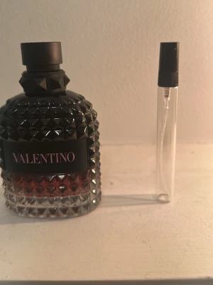 Valentino parfym born in Roma intense - Snygg parfymflaska från Valentino med svart och transparent glas, dekorerad med nitar för en edgy look. Flaskan har en svart etikett med rosa text och kommer med en liten genomskinlig sprayflaska. Perfekt för dig som gillar lyxiga dofter.