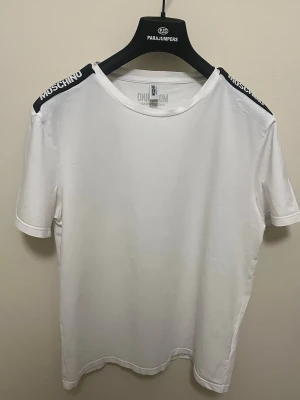 Vit t-shirt från Moschino - Snygg vit t-shirt från Moschino med svarta axelband där märkesnamnet står i vitt. Klassisk rund halsringning och korta ärmar. Tillverkad i mjuk bomull som känns skön mot huden. Perfekt för en clean och stilren look.