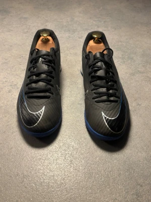 Nike Mercurial svart/blå fotbollsskor - Snygga Nike Mercurial futsal skor i svart med blå detaljer och silverfärgad swoosh. Skorna har snörning och är designade för inomhusspel. Lätta och smidiga med syntetmaterial för bästa bollkontroll.