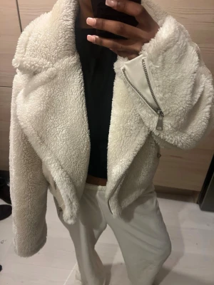 Vit croppad teddyjacka NaKd - Säljer en supermjuk och fluffig vit teddyjacka i croppad modell. Jackan har bred krage, långa ärmar och coola dragkedjedetaljer. Perfekt för dig som vill ha en trendig och cozy look till hösten eller vintern.