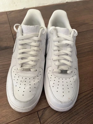 Nike Air Force 1 vita sneakers - Fräscha Nike Air Force 1 sneakers i helvitt skinn med klassisk rund tå och platt sula. Snygga detaljer som perforeringar på tån och metallbricka med 'AF1' på snörningen. Perfekt för dig som gillar cleana och tidlösa sneakers med streetkänsla.